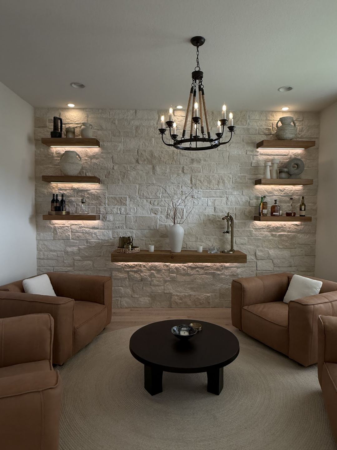 Home lounge with a stone accent wall, floating shelves lit from below, recessed ceiling lights and a chandelier